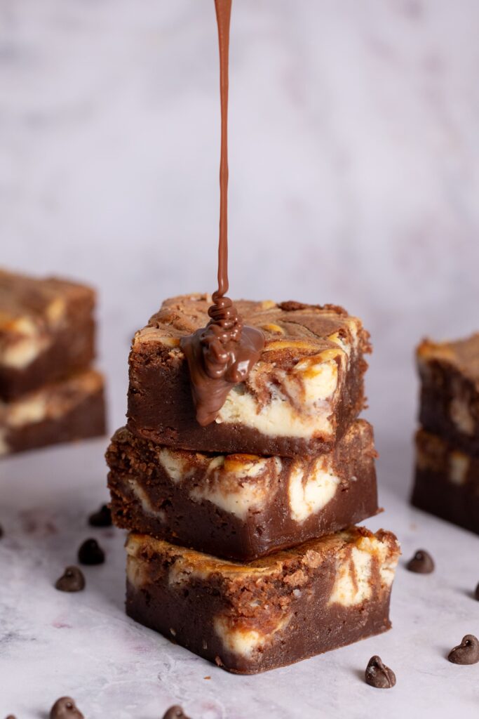 Pila de tres brownies marmolados sobre una superficie gris con un chorrito de salsa de chocolate cayendo desde arriba. Hay pepitas de chocolate esparcidas por todas partes y se ve otro brownie en el fondo.
