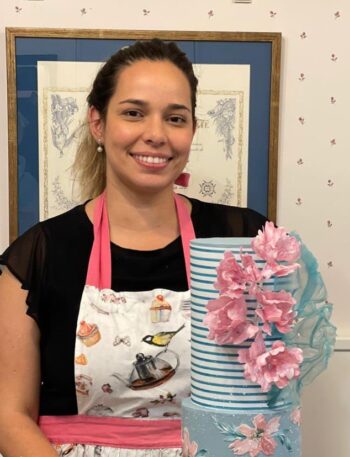 Una mujer sonríe, de pie junto a un pastel alto de rayas azules adornado con flores rosas. Lleva un delantal con diseños coloridos, y al fondo se ven obras de arte enmarcadas y una pared con patrones sutiles.