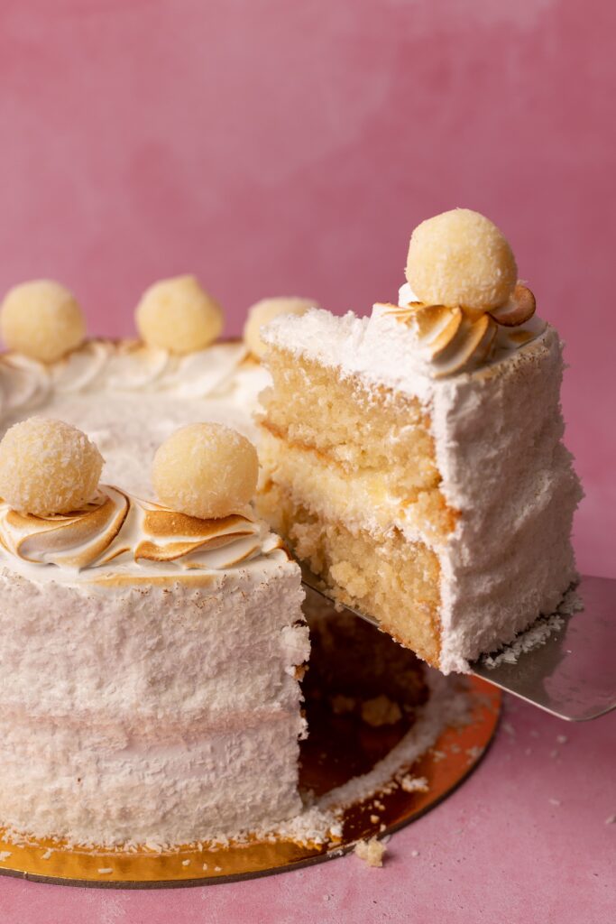 Se saca una porción de pastel de coco con glaseado de merengue con varias capas. El pastel está cubierto con trufas redondas de coco y espirales de glaseado. El fondo es rosa.