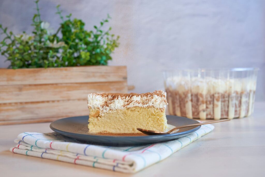 Una porción de pastel de tres leches cubierto con crema batida y canela en un plato con una cuchara. Reposa sobre un mantel a cuadros sobre una mesa. En el fondo, una caja de madera contiene plantas, junto a tazas transparentes llenas de postres en capas.