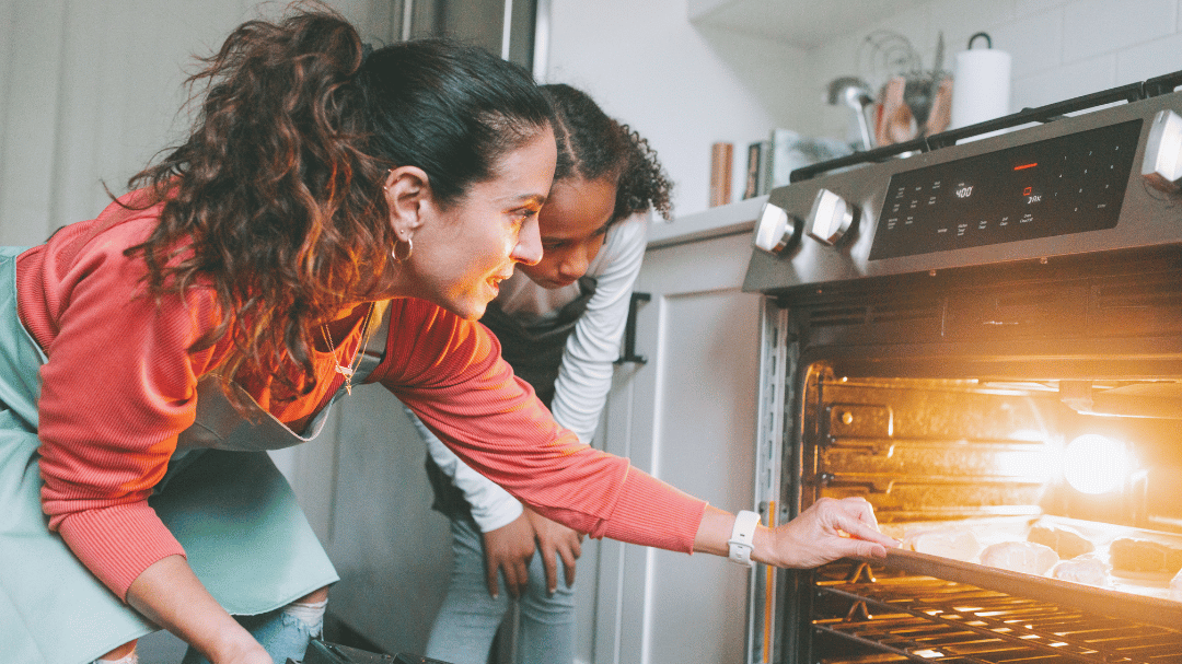 Dos personas en una cocina supervisan la cocción de los alimentos en un horno. Una, con cola de caballo, camisa roja y delantal, se agacha para mirar dentro. La otra, de pelo rizado, observa atentamente. La cocina está tenuemente iluminada y es acogedora.