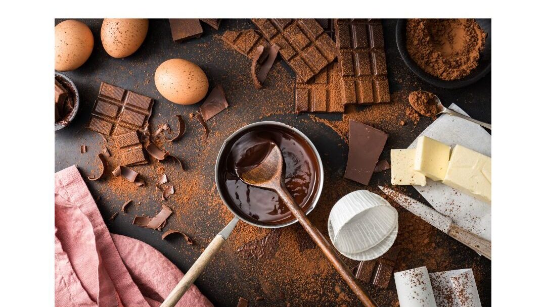 Una disposición plana de ingredientes para hornear sobre una mesa, incluyendo cuadrados de chocolate, huevos, un tazón de chocolate derretido, cacao en polvo, mantequilla y un batidor. También hay un paño rosa a un lado, creando una cálida y acogedora escena de repostería.