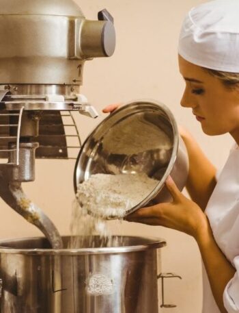 Un panadero con uniforme blanco y gorro de chef vierte harina de un recipiente metálico en una gran batidora industrial en una panadería. La batidora es de acero inoxidable y el ambiente es limpio y profesional.
