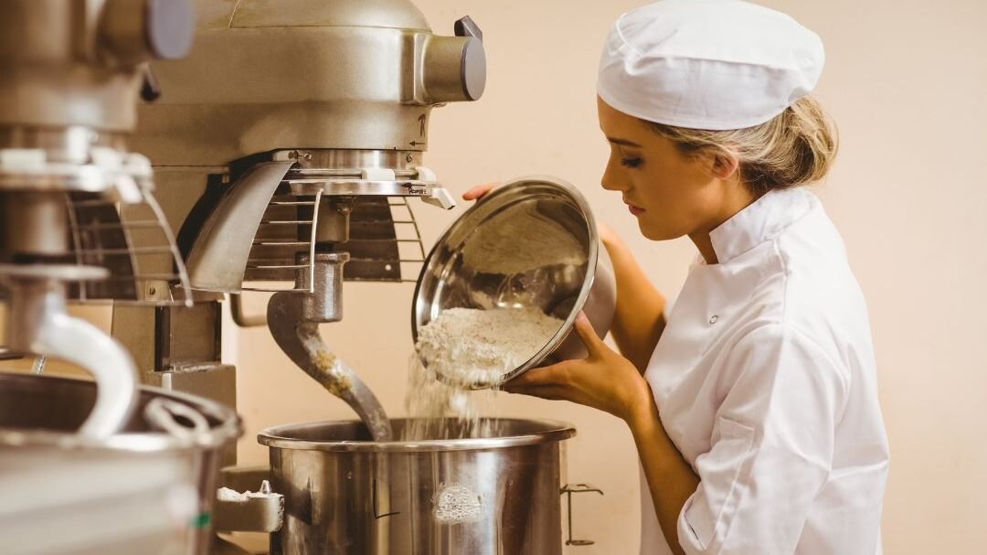 Un panadero con uniforme blanco y gorro de chef vierte harina de un recipiente metálico en una gran batidora industrial en una panadería. La batidora es de acero inoxidable y el ambiente es limpio y profesional.