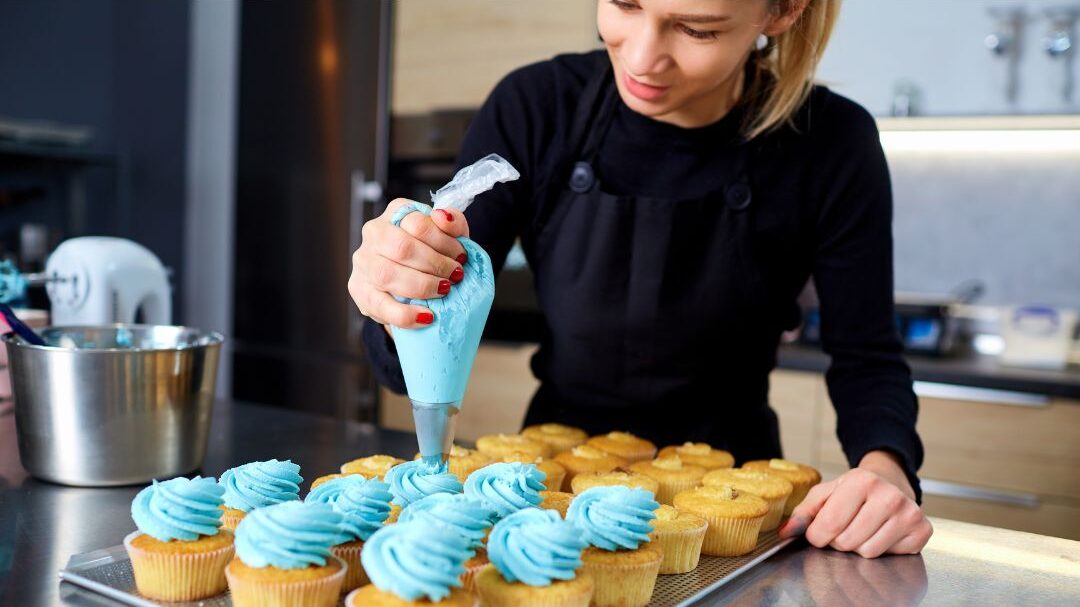 Una persona con un delantal negro decora cupcakes con glaseado azul en una cocina. Sostiene una manga pastelera y añade espirales de glaseado a la superficie. Al fondo se ven una bandeja de cupcakes sencillos y una batidora.