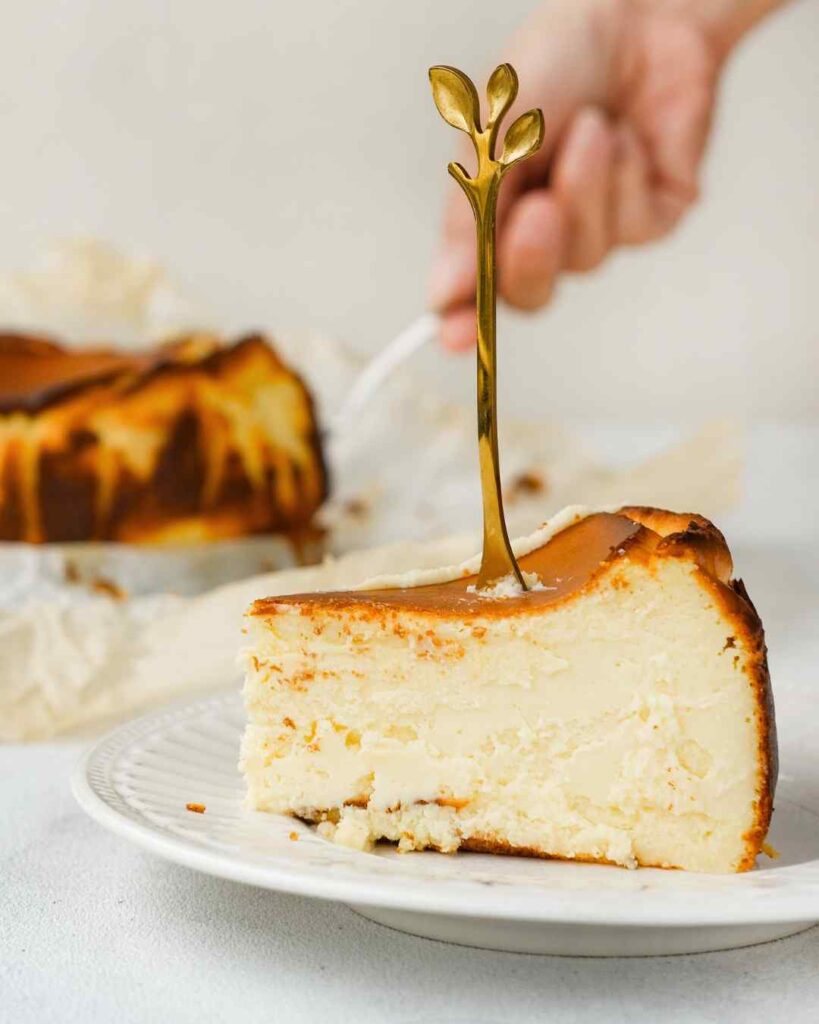 Una porción de tarta de queso cremosa con un tenedor dorado sobre un plato blanco. Una mano sostiene un tenedor en el fondo, cerca del resto de la tarta de queso. La tarta de queso de la viña tiene una superficie ligeramente dorada y una textura suave. El fondo está difuminado y de color claro.