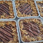 Bandejas de aluminio llenas de marquesas denutella en capas, cubiertos con llovizna de chocolate y galletas trituradas. Cada bandeja muestra un patrón similar y está dispuesta en filas muy juntas.