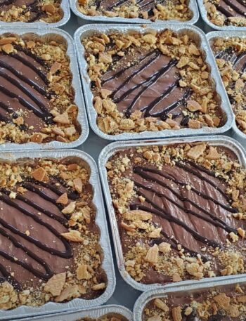 Bandejas de aluminio llenas de marquesas denutella en capas, cubiertos con llovizna de chocolate y galletas trituradas. Cada bandeja muestra un patrón similar y está dispuesta en filas muy juntas.