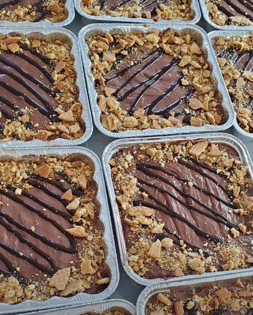 Bandejas de aluminio llenas de marquesas denutella en capas, cubiertos con llovizna de chocolate y galletas trituradas. Cada bandeja muestra un patrón similar y está dispuesta en filas muy juntas.