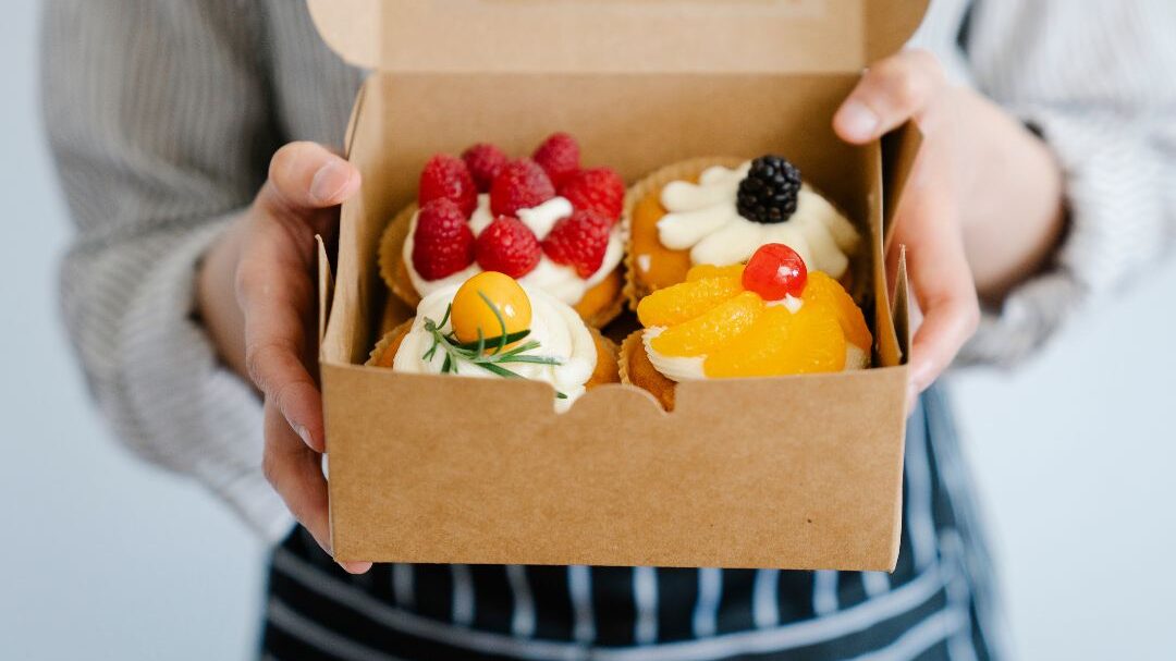 Una persona sostiene una caja de cartón abierta con cuatro cupcakes surtidos, cada uno decorado con frutas como frambuesas, una mora, un physalis y una rodaja de naranja. Lleva un delantal a rayas y una camisa a rayas.